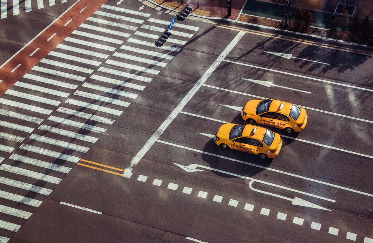 hero-img-01 Two yellow taxis navigate a city intersection with marked pedestrian crossings.